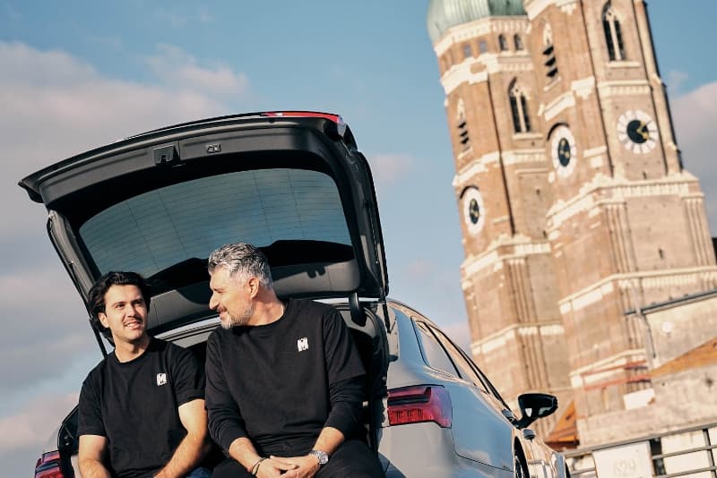 Das Mosaik Motos Team sitzend und lächelnd im offenen Kofferraum eines ihrer hochwertigen Audis vor der Frauenkirche in München
